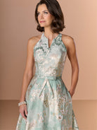 Woman wearing a floral dress against a beige background / This stunning gown features a softly muted brocade fabric with a halter neckline and a belted waist.