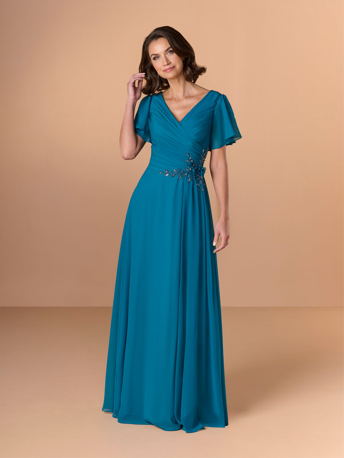 A woman modeling a floor-length teal blue gown with short flutter sleeves, a V-neckline, and a beaded embellishment at the waist, set against a solid beige studio background. / A-line chiffon style with V-neck and short flatter sleeves. Finely gathering on bodice finished with hand beading motif on side waist area.
