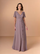 A woman modeling a floor-length mauve-gray evening gown with short flutter sleeves, a V-neckline, and a decorative embellished waistband, set against a solid warm beige studio backdrop. / A-line chiffon style with V-neck and short flatter sleeves. Finely gathering on bodice finished with hand beading motif on side waist area.