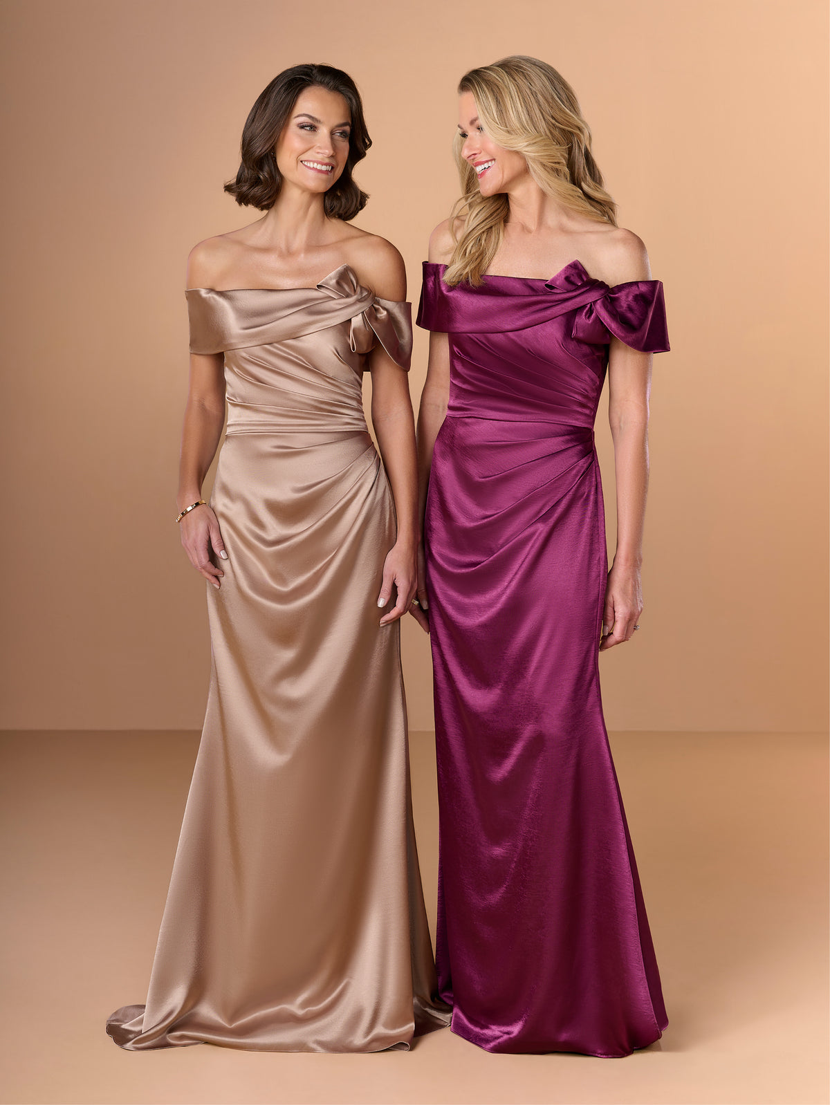 Two women modeling off-the-shoulder satin floor-length dresses against a warm beige studio background. The woman on the left wears a champagne beige dress, and the woman on the right wears a rich magenta-purple dress. / Slim charmeuse style with straight neckline and off-shoulders collar with twist and bow on side front. Pleating on front dress.