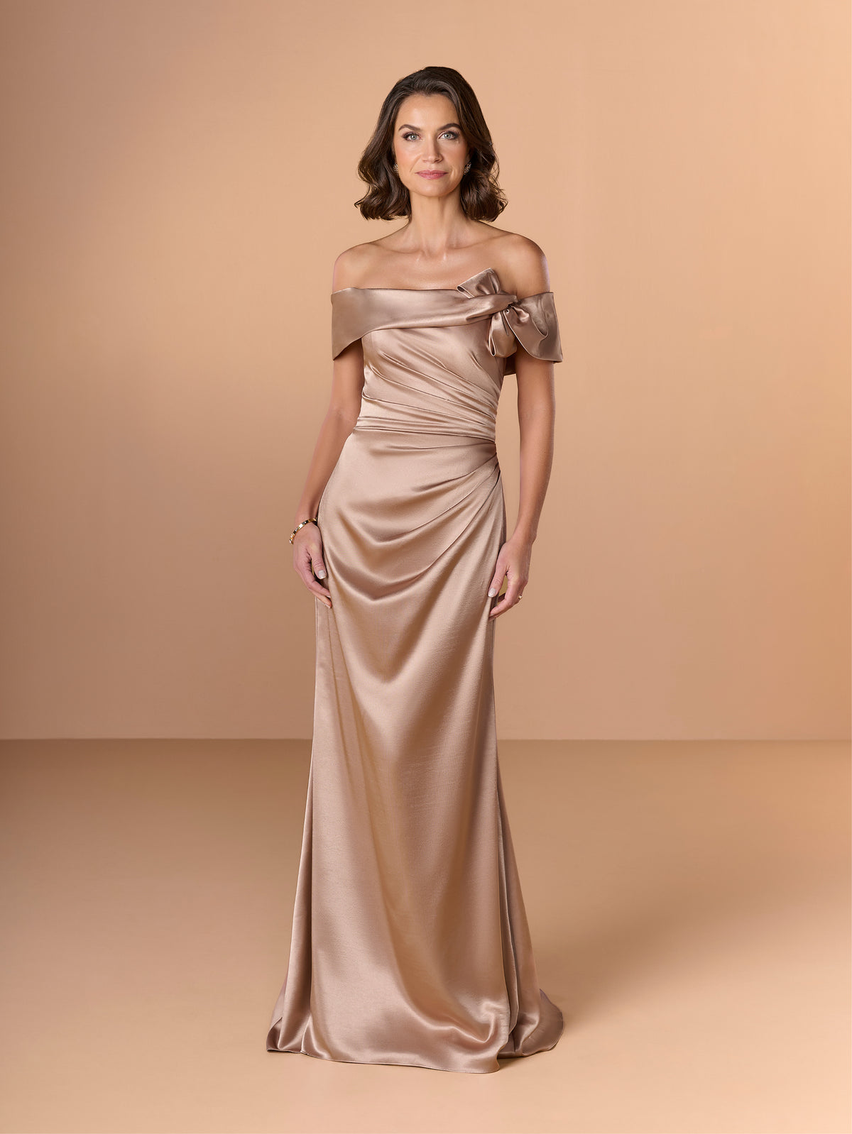 A woman modeling a floor-length off-the-shoulder satin gown in a champagne beige color, standing against a solid warm beige studio backdrop. / Slim charmeuse style with straight neckline and off-shoulders collar with twist and bow on side front. Pleating on front dress.
