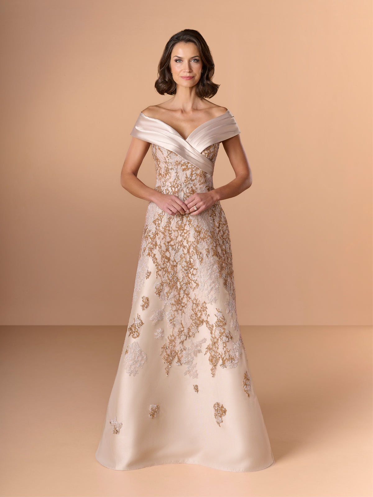 A woman models an elegant off-the-shoulder evening gown in champagne beige with intricate gold floral embroidery, set against a solid warm beige studio background. / A-line lace dress with mikado collar off-shoulder.