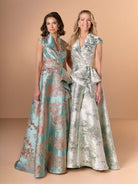 Two women modeling formal evening gowns against a warm beige studio backdrop. The gown on the left is sleeveless with a wrap-style bodice and a flowing skirt in aqua with pink floral patterning. / A-line style in brocade with V-neck and standing collar at the back neckline. Side front pleating on bodice finished with short ruffle.