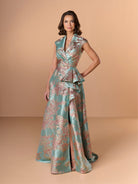 Woman wearing a floral patterned dress on a beige background / A-line style in brocade with V-neck and standing collar at the back neckline. Side front pleating on bodice finished with short ruffle.