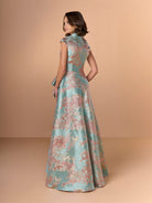 Woman wearing a floral evening gown against a beige background / A-line style in brocade with V-neck and standing collar at the back neckline. Side front pleating on bodice finished with short ruffle.