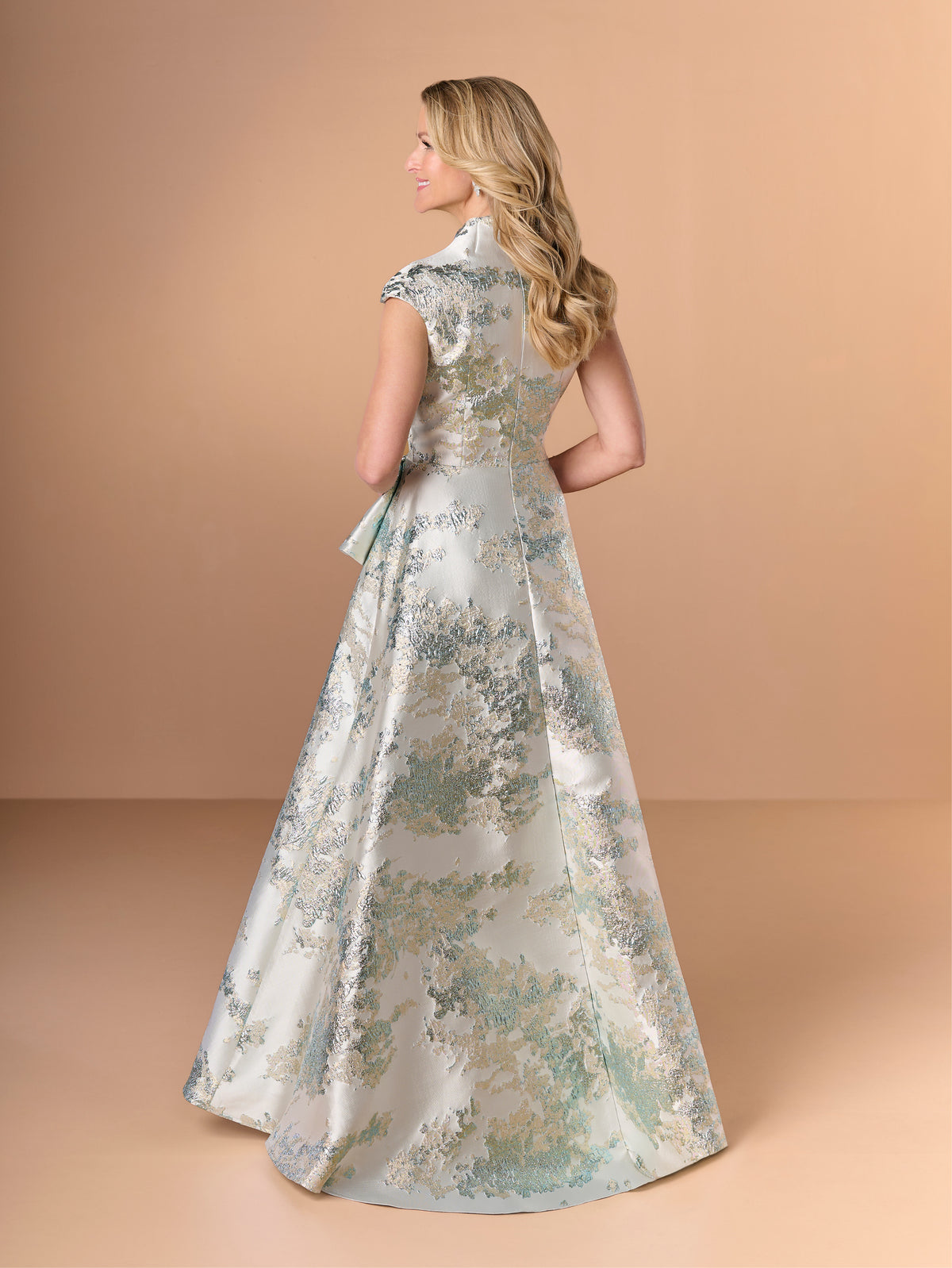 A woman modeling a floor-length formal gown from the back, shown against a warm beige studio backdrop. The dress has a shimmering light silver base with an allover floral jacquard pattern in soft blue, sage green, and beige tones. / A-line style in brocade with V-neck and standing collar at the back neckline. Side front pleating on bodice finished with short ruffle.