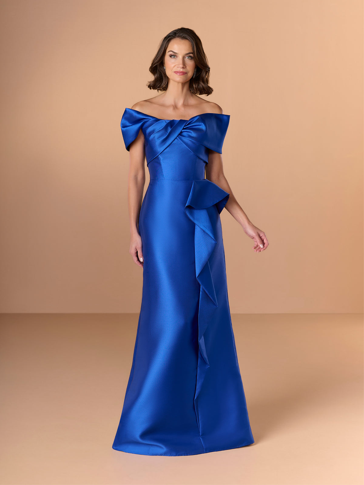 A woman modeling an off-the-shoulder floor-length gown in rich royal blue satin, set against a solid warm beige studio background. / Slim mikado style with straight neckline and off-shoulder straps. Draping on bust area and frill feature on side front skirt.