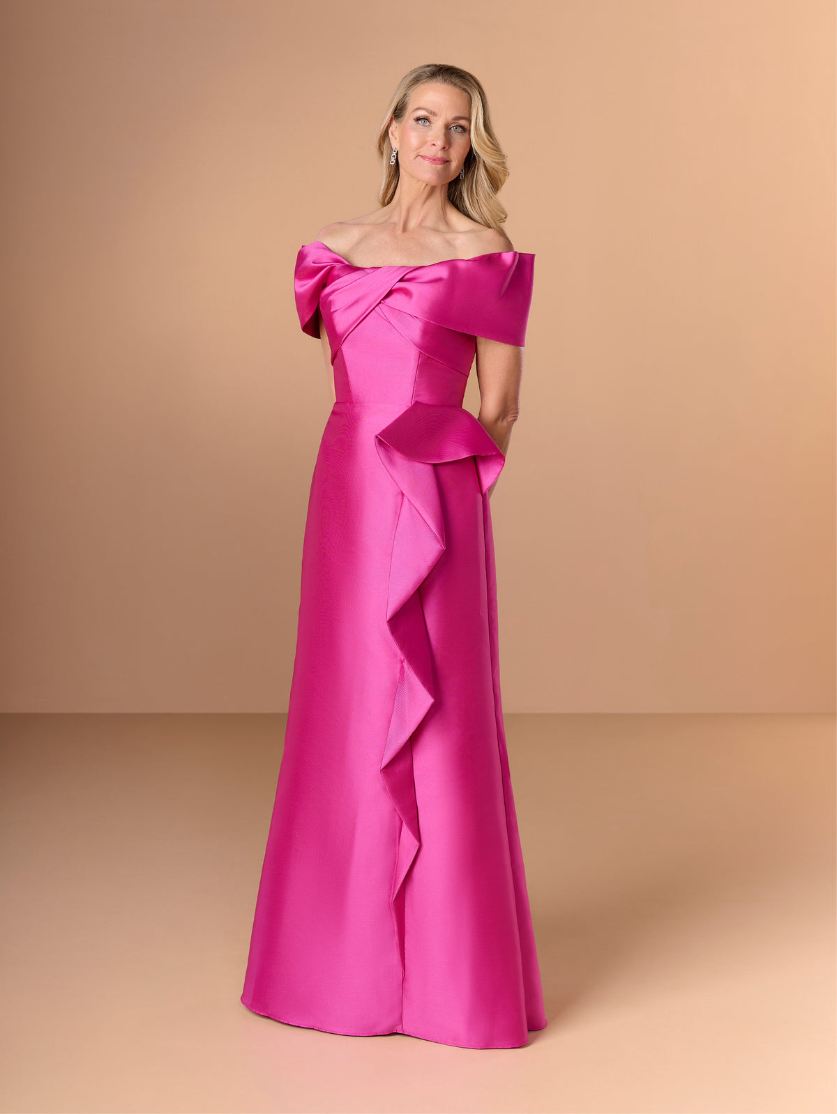 A woman modeling a bright fuchsia off-the-shoulder floor-length gown with a large bow at the neckline and cascading ruffle detailing down the front, set against a solid beige studio backdrop. / Slim mikado style with straight neckline and off-shoulder straps. Draping on bust area and frill feature on side front skirt.