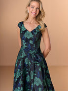 Woman wearing a dark blue and green floral dress against a beige background / A-line brocade style with V-neck and short sleeves. Waist band draping with soft bow on side. Invisible zipper closure at the back.