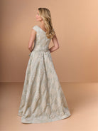 A woman modeling a floor-length A-line gown from the back, featuring a textured jacquard pattern in beige and muted sage green on a warm peach studio background. / A-line brocade style with V-neck and short sleeves. Waist band draping with soft bow on side. Invisible zipper closure at the back.