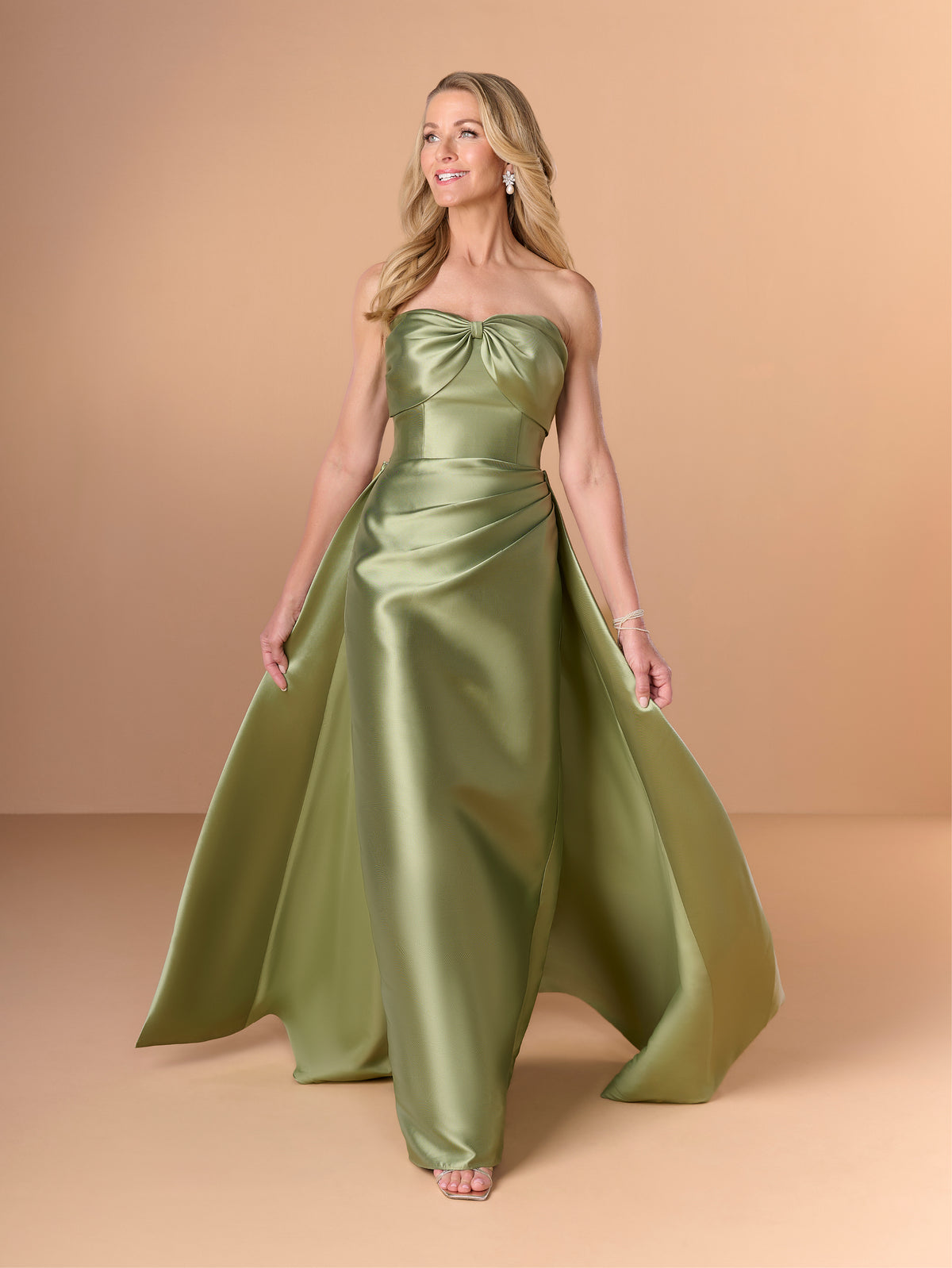 A woman modeling a strapless floor-length satin gown in a muted olive green color, set against a solid warm beige studio background. / Slim mikado style with draping on bust area in bow shape going around to the back. Side front draping on stomach area. Detachable back skirt sweep.