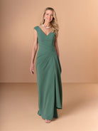 A woman modeling a floor-length sage green gown with a V-neckline, cap sleeves, and a draped gathered waist, photographed against a warm beige studio backdrop. / Slim crepe style with V-neck and cup shoulder. Side pleating on front dress with long ruffle on side.