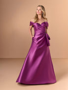 A woman modeling an off-the-shoulder satin formal gown in a rich magenta-purple color, standing against a solid warm beige studio backdrop. / A-line mikado style with sweetheart neckline and off-shoulder straps, front draping with bow feature on side.