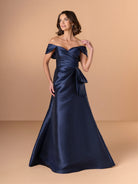 A woman modeling an off-the-shoulder navy blue satin evening gown with a sweetheart neckline, gathered waist, and flared skirt against a warm beige studio backdrop. / A-line mikado style with sweetheart neckline and off-shoulder straps, front draping with bow feature on side.