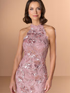 A woman modeling a sleeveless, high-neck, floor-length evening gown in dusty pink with intricate sequined floral and leaf patterns, set against a solid warm beige studio background. / This dress is a fitted, floor-length gown featuring a high halter neckline and an all-over floral pattern made of shimmering sequins and embroidery. The design follows a sleek, straight silhouette that highlights the embellished details from top to bottom, creating an elegant and refined look.