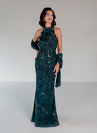 A woman modeling a floor-length dark teal evening gown with intricate floral beadwork and sequins, paired with matching sheer draped gloves, against a neutral light gray studio background. / This dress is a fitted, floor-length gown featuring a high halter neckline and an all-over floral pattern made of shimmering sequins and embroidery. The design follows a sleek, straight silhouette that highlights the embellished details from top to bottom, creating an elegant and refined look.