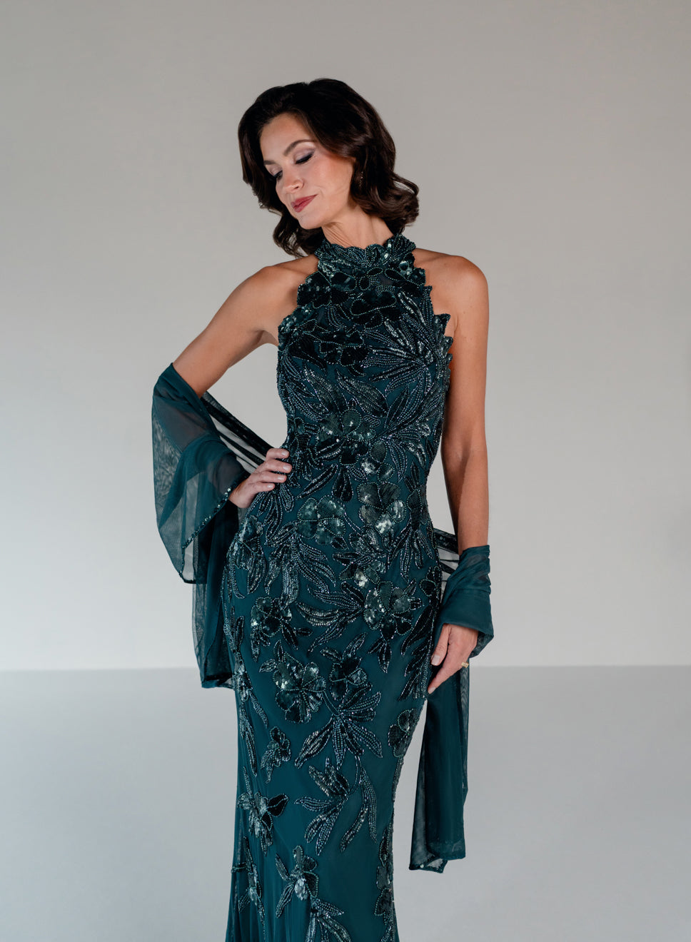 A woman models a dark teal halter-neck evening gown with intricate sequin and bead floral detailing, paired with matching sheer draped gloves, against a neutral studio background. / This dress is a fitted, floor-length gown featuring a high halter neckline and an all-over floral pattern made of shimmering sequins and embroidery. The design follows a sleek, straight silhouette that highlights the embellished details from top to bottom, creating an elegant and refined look.