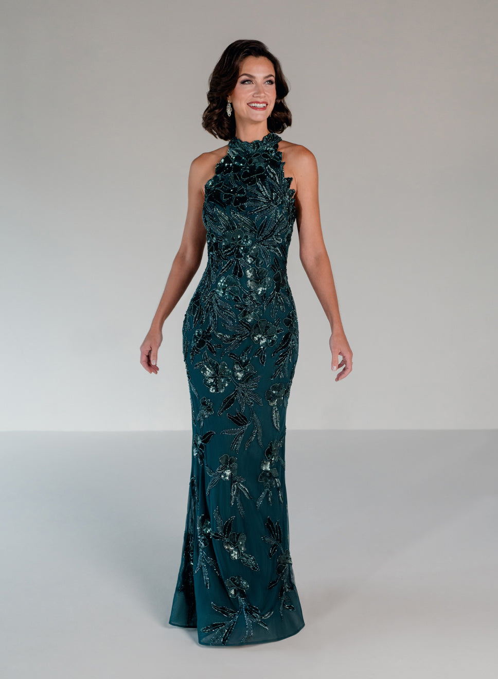 A woman modeling a floor-length, halter-neck evening gown in deep teal with silver and black floral embroidery, set against a neutral light gray studio background. / This dress is a fitted, floor-length gown featuring a high halter neckline and an all-over floral pattern made of shimmering sequins and embroidery. The design follows a sleek, straight silhouette that highlights the embellished details from top to bottom, creating an elegant and refined look.
