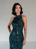 A woman modeling a sleeveless, high-neck, floor-length gown in deep teal with intricate floral applique and sequined detailing, set against a neutral light gray studio background. / This dress is a fitted, floor-length gown featuring a high halter neckline and an all-over floral pattern made of shimmering sequins and embroidery. The design follows a sleek, straight silhouette that highlights the embellished details from top to bottom, creating an elegant and refined look.