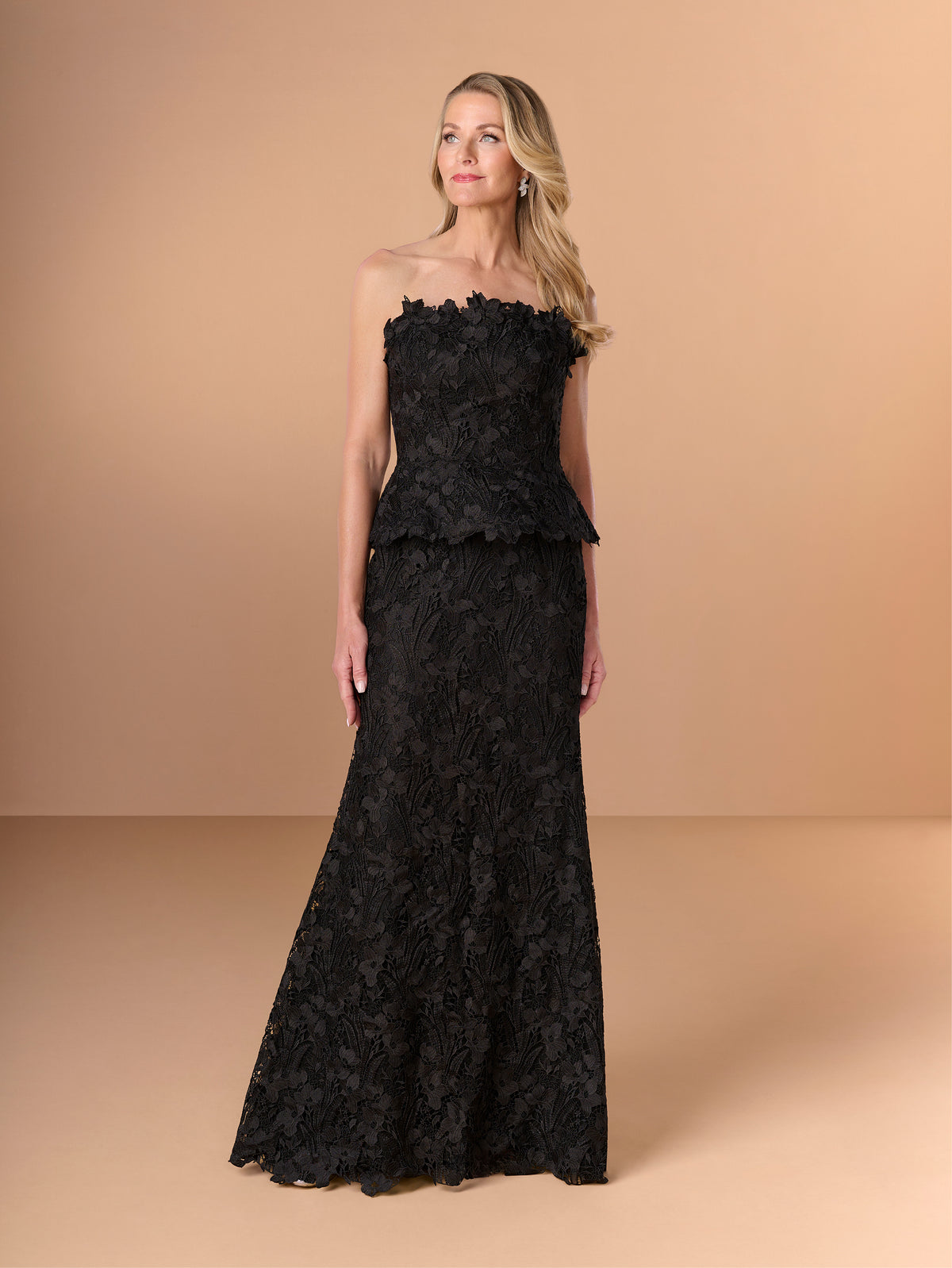 A woman modeling a strapless black lace formal gown with a ruffled peplum waist, photographed against a warm beige studio backdrop. / Created with a truly exquisite lace, this strapless gown features a figure-flattering peplum with a stand-away organically cut neckline.