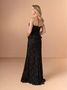 A woman modeling a strapless black lace evening gown from the back, standing against a solid warm beige studio backdrop. / Created with a truly exquisite lace, this strapless gown features a figure-flattering peplum with a stand-away organically cut neckline.