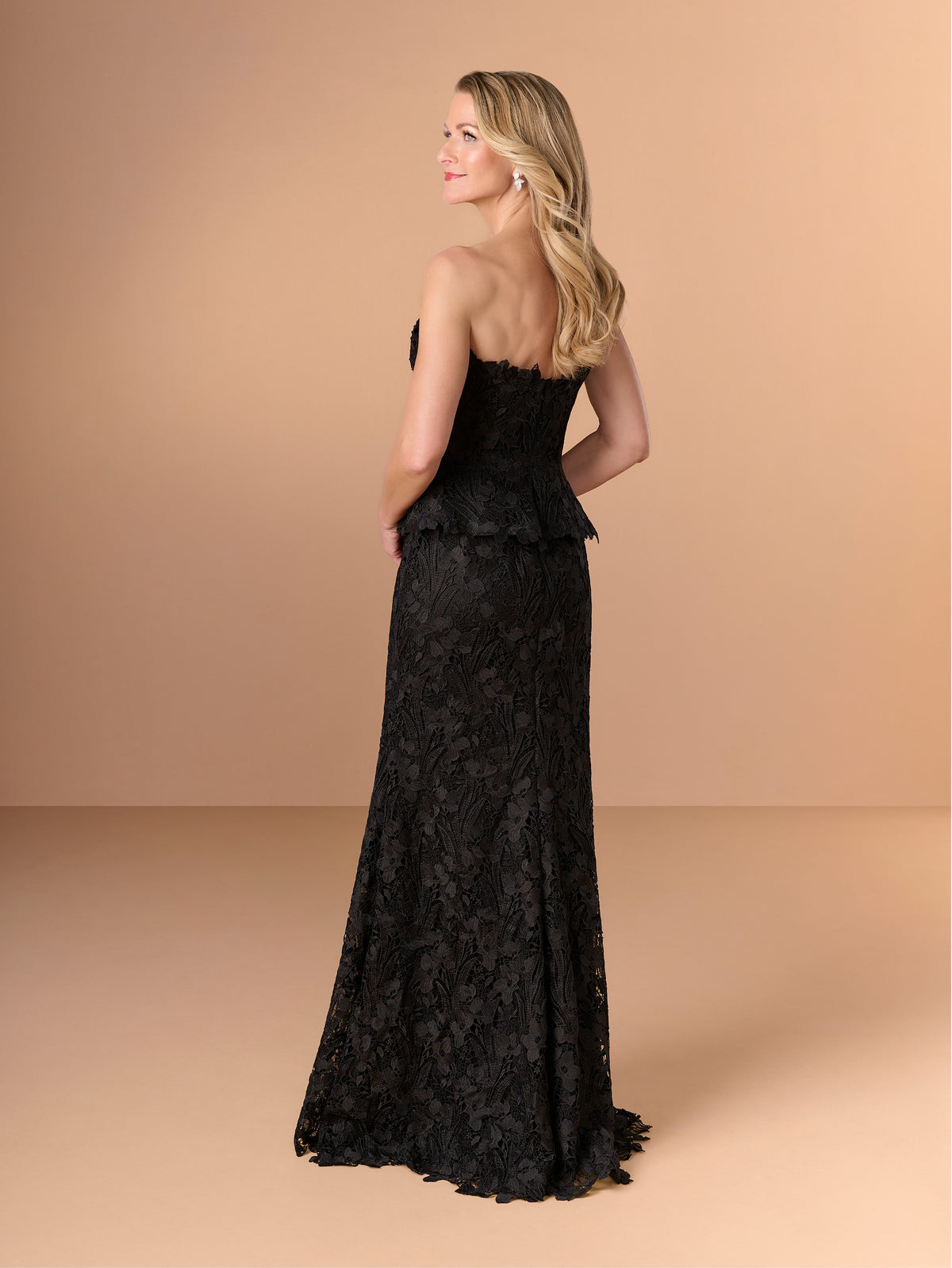 A woman modeling a strapless black lace evening gown from the back, standing against a solid warm beige studio backdrop. / Created with a truly exquisite lace, this strapless gown features a figure-flattering peplum with a stand-away organically cut neckline.