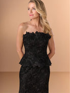 A blonde woman wearing a strapless black lace evening gown with a textured floral applique bodice and peplum, posed against a solid beige studio background. / Created with a truly exquisite lace, this strapless gown features a figure-flattering peplum with a stand-away organically cut neckline.