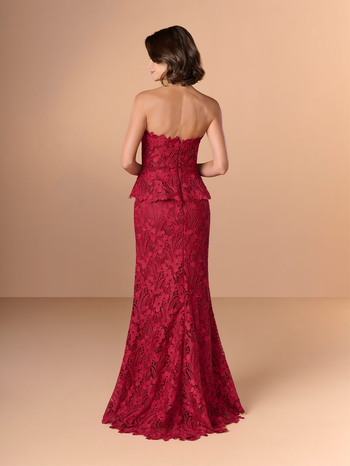 Woman wearing a red lace evening gown against a beige background / Created with a truly exquisite lace, this strapless gown features a figure-flattering peplum with a stand-away organically cut neckline.