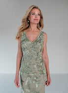 A woman modeling a sleeveless V-neck evening gown with metallic sage green sequin leaf embroidery over a taupe-beige mesh base, set against a neutral gray studio background. / This gown features a sleeveless, fitted silhouette with a softly draped v-neckline. The entire dress is decorated with shimmering floral and leaf-patterned embellishments that extend from top to hem. Its sleek, floor-length design creates an elegant and refined look from both the front and back.