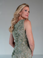 A blonde woman wearing a sleeveless, one-shoulder embellished dress in a shimmering sage green and silver sequin pattern, photographed against a neutral gray studio background. / This gown features a sleeveless, fitted silhouette with a softly draped v-neckline. The entire dress is decorated with shimmering floral and leaf-patterned embellishments that extend from top to hem. Its sleek, floor-length design creates an elegant and refined look from both the front and back.