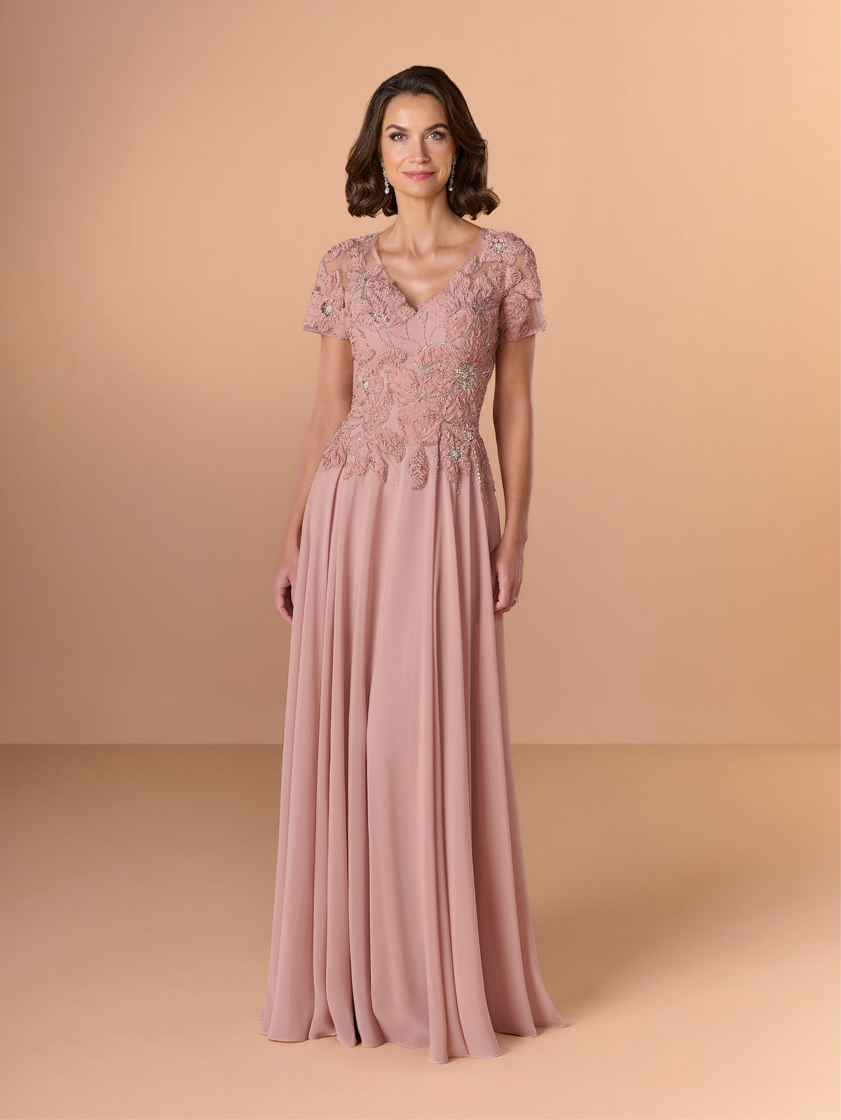 A woman modeling a floor-length dusty pink evening gown with short lace sleeves, a V-neckline, and intricate floral lace applique across the bodice, set against a solid warm beige studio background. / A-line chiffon style with bodice coved with beaded motifs, V-neck short semi sheer sleeves.