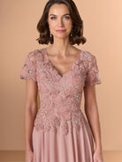 A woman wearing a dusty pink evening gown with intricate floral lace embroidery, short sleeves, and a V-neckline, set against a solid beige studio background. / A-line chiffon style with bodice coved with beaded motifs, V-neck short semi sheer sleeves.