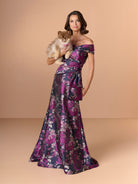 Woman in a purple floral dress holding a small dog against a beige background / Slim brocade style with collar/straps twist, pleating on front bodice.