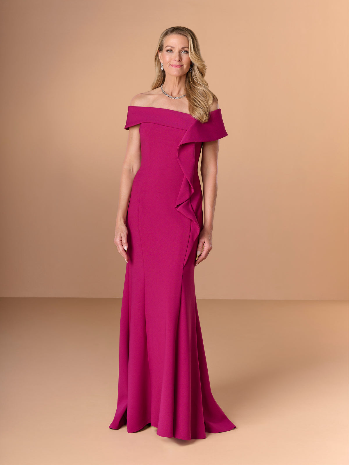 A woman modeling a bright magenta off-the-shoulder evening gown with a ruffled asymmetrical detail on the bodice and skirt, set against a solid warm beige studio background. / Slim crepe style with asymmetrical wide collar and side raffle.