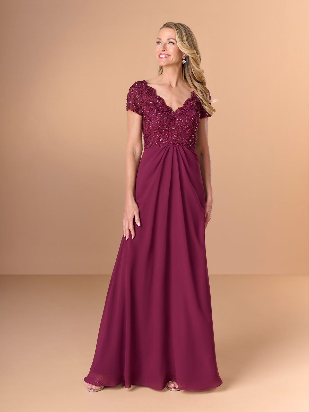 A woman modeling a floor-length burgundy formal gown with a beaded lace bodice, short cap sleeves, and a flowing skirt against a solid tan studio background. / A-line style with lace bodice and chiffon skirt. V-neck edge and short sleeves hemline edge finished with scallops. Draping on front and back skirt in fan shape.