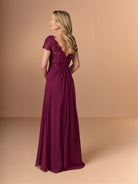 A woman modeling a floor-length burgundy evening gown from the back, featuring a beaded lace bodice, short cap sleeves, and a flowing skirt, set against a solid beige studio background. / A-line style with lace bodice and chiffon skirt. V-neck edge and short sleeves hemline edge finished with scallops. Draping on front and back skirt in fan shape.