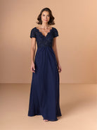 A woman modeling a navy blue floor-length gown with a beaded, scalloped sweetheart bodice and short cap sleeves, set against a solid beige studio backdrop. / A-line style with lace bodice and chiffon skirt. V-neck edge and short sleeves hemline edge finished with scallops. Draping on front and back skirt in fan shape.