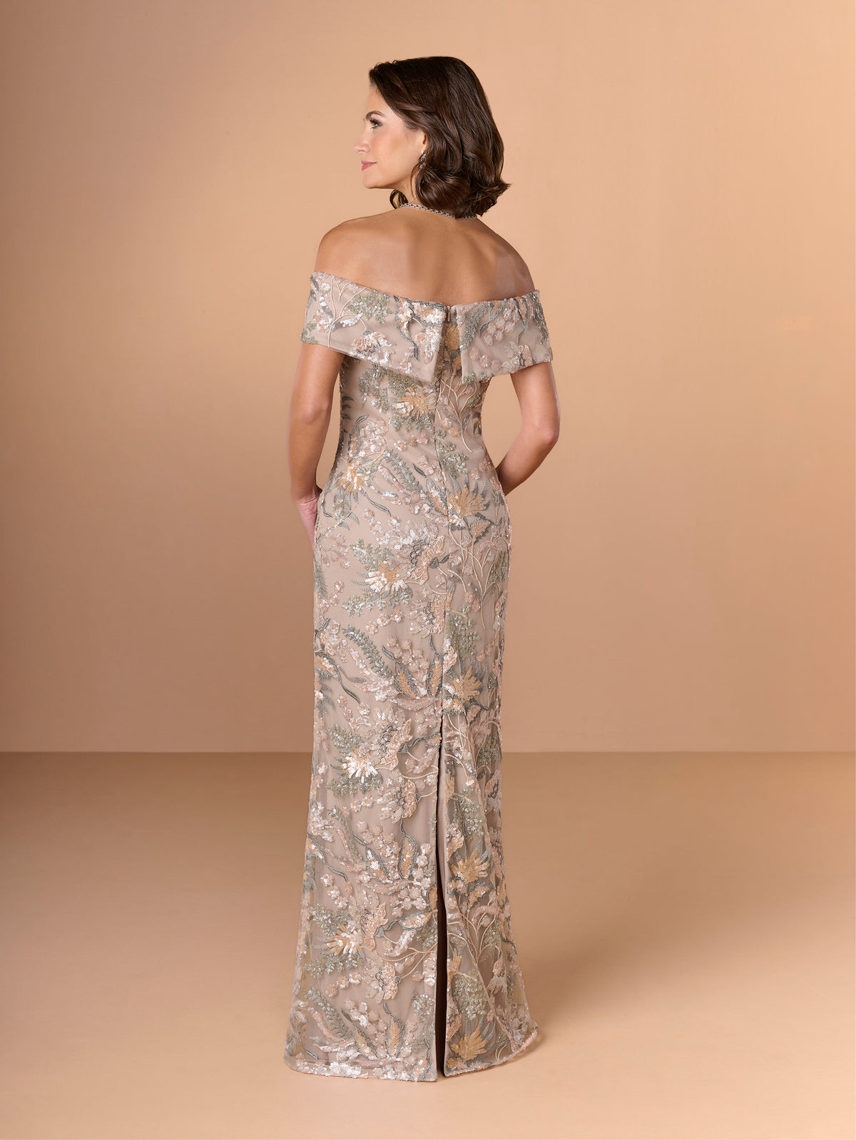 A woman modeling an off-the-shoulder floor-length gown from the back, featuring a shimmering metallic taupe and gold floral pattern against a solid warm beige studio backdrop. / Slim lace style with straight neckline and wide off shoulder collar. Split on center back skirt.