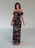 A woman modeling an off-the-shoulder floor-length gown with a black base and an intricate multicolor floral and vine pattern in pink, orange, and white, set against a plain light gray studio background. / Slim lace style with straight neckline and wide off shoulder collar. Split on center back skirt.