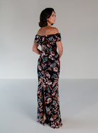 A woman modeling an off-the-shoulder black maxi dress with a vibrant pink, orange, and green floral and bird pattern, photographed against a neutral light gray studio background. / Slim lace style with straight neckline and wide off shoulder collar. Split on center back skirt.