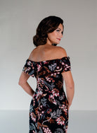A woman wearing an off-the-shoulder black dress with a dense pink, white, and brown floral print, photographed from a three-quarters back angle against a neutral light gray studio background. / Slim lace style with straight neckline and wide off shoulder collar. Split on center back skirt.