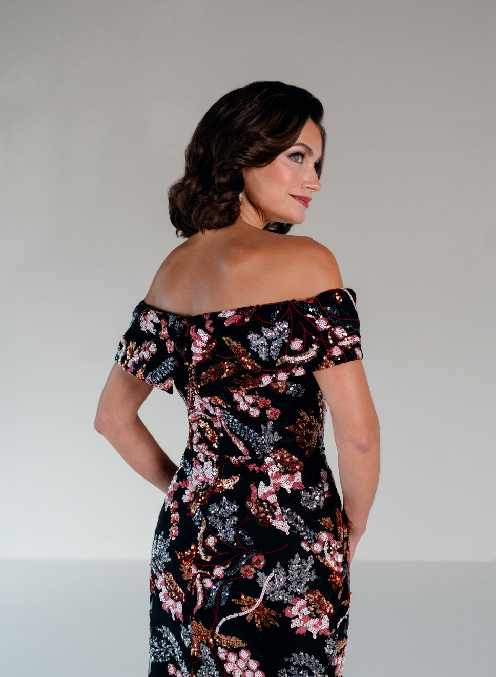 A woman wearing an off-the-shoulder black dress with a dense pink, white, and brown floral print, photographed from a three-quarters back angle against a neutral light gray studio background. / Slim lace style with straight neckline and wide off shoulder collar. Split on center back skirt.