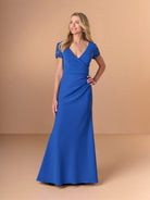 A woman modeling a floor-length royal blue gown with short lace cap sleeves, a V-neckline, and a fitted mermaid silhouette against a warm beige studio backdrop. / Short sleeve gown with beaded sleeve and body pleating detail.