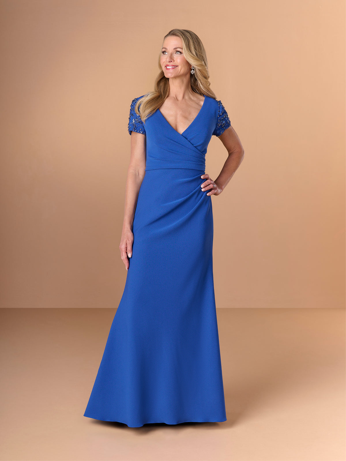 A woman modeling a floor-length royal blue gown with short lace cap sleeves, a V-neckline, and a fitted mermaid silhouette against a warm beige studio backdrop. / Short sleeve gown with beaded sleeve and body pleating detail.