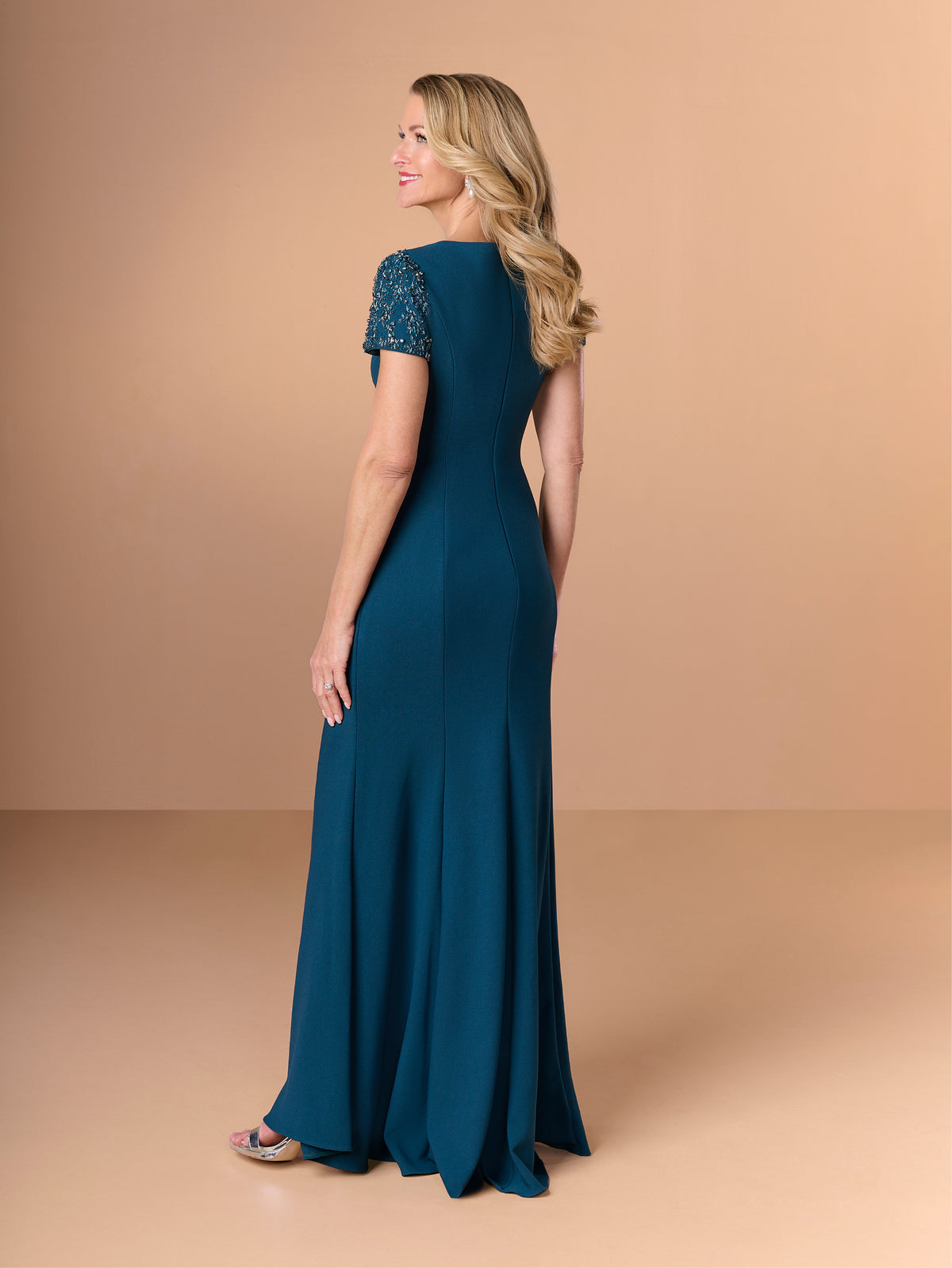 Woman wearing a teal evening gown with lace sleeves on a beige background / Short sleeve gown with beaded sleeve and body pleating detail.