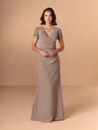 A woman modeling a floor-length taupe gown with lace short sleeves, a V-neckline, and a draped wrap-style bodice, photographed against a warm beige studio backdrop. / Short sleeve gown with beaded sleeve and body pleating detail.