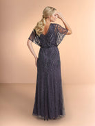 A woman modeling a floor-length evening gown from the back, featuring sheer lace short sleeves, a V-neckline, and intricate bead and sequin embellishments on a dark gray tulle skirt, set against a solid peach studio backdrop. / All over beaded gown with v neckline and long flutter sleeve.