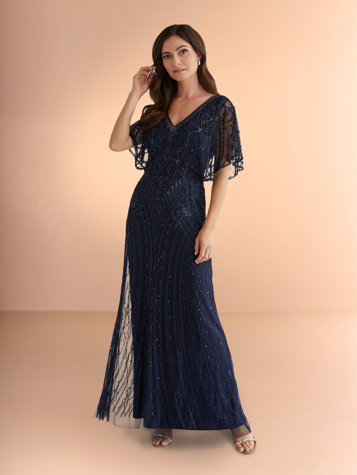 Woman wearing a navy blue evening gown with sheer sleeves against a beige background / All over beaded gown with v neckline and long flutter sleeve.