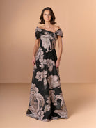 A woman modeling an off-the-shoulder black maxi dress with a large beige floral print, set against a solid beige studio background. / A-line style in textured organza with asymmetrical neckline and pleated wide collar off-shoulder.