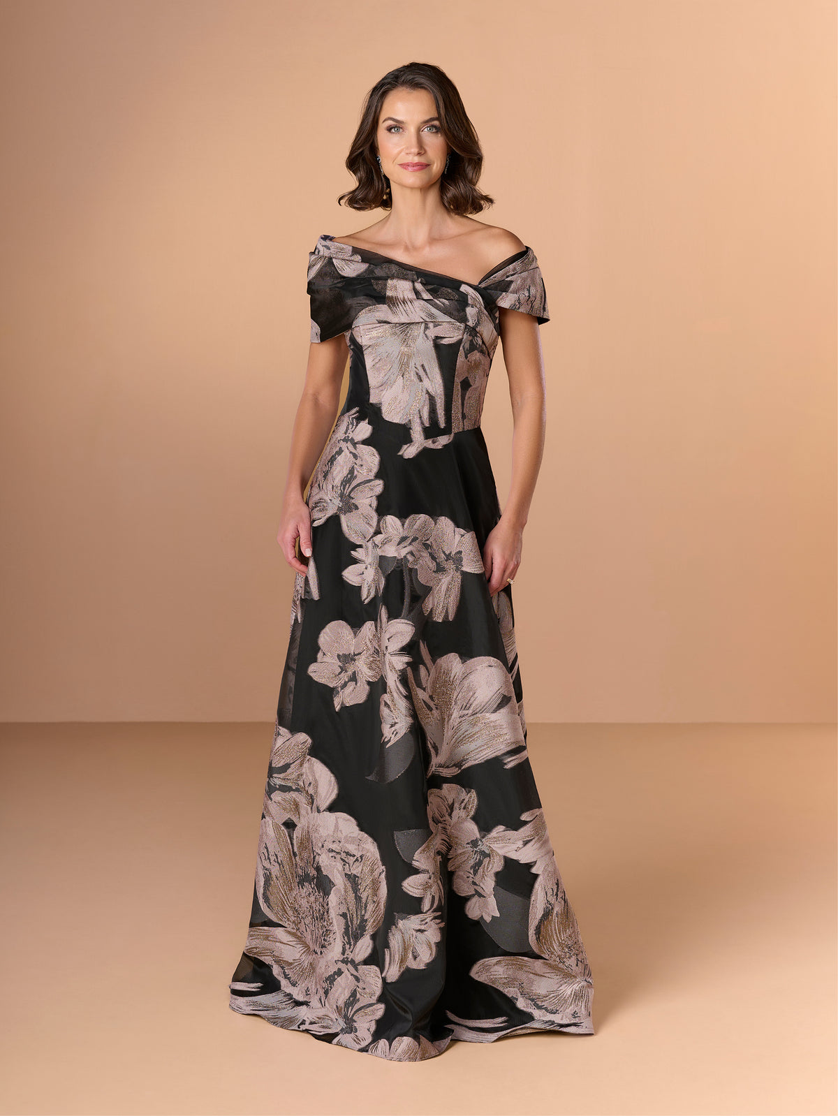 A woman modeling an off-the-shoulder black maxi dress with a large beige floral print, set against a solid beige studio background. / A-line style in textured organza with asymmetrical neckline and pleated wide collar off-shoulder.